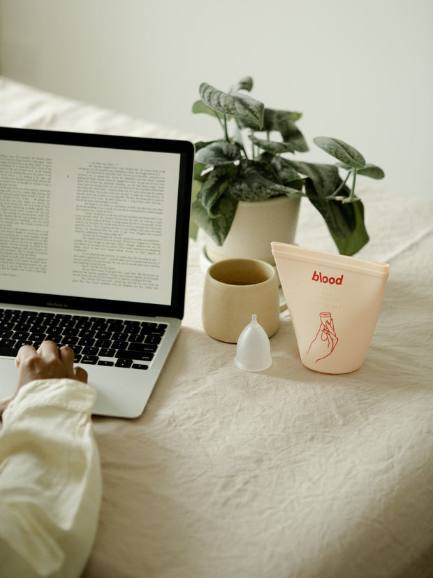 a girl is sitting at a table using her laptop. there is a menstrual cup and sanitising pouch on the table.